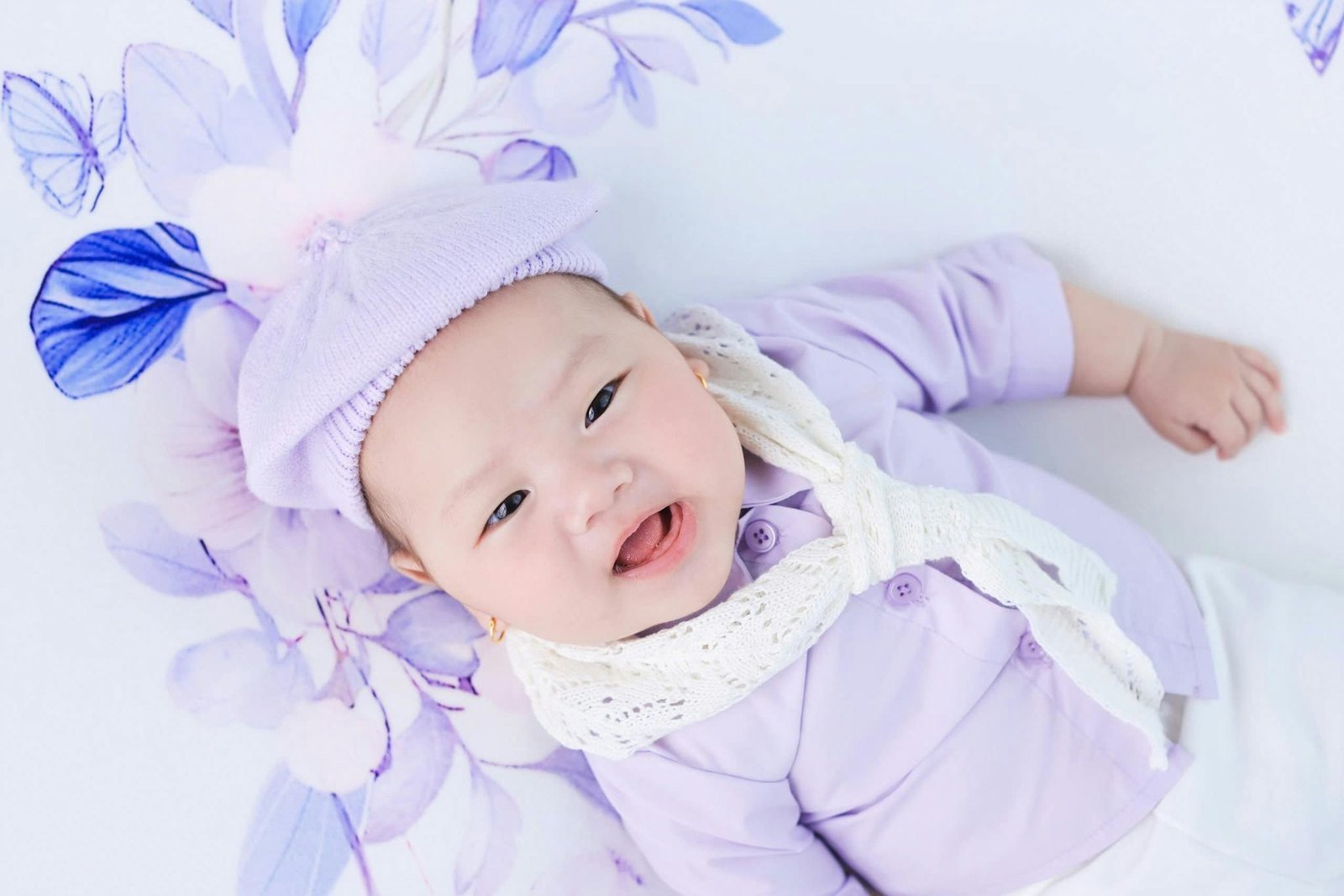 A cute baby in lavender attire, wearing a knit beret and white scarf, lying on a floral background.
