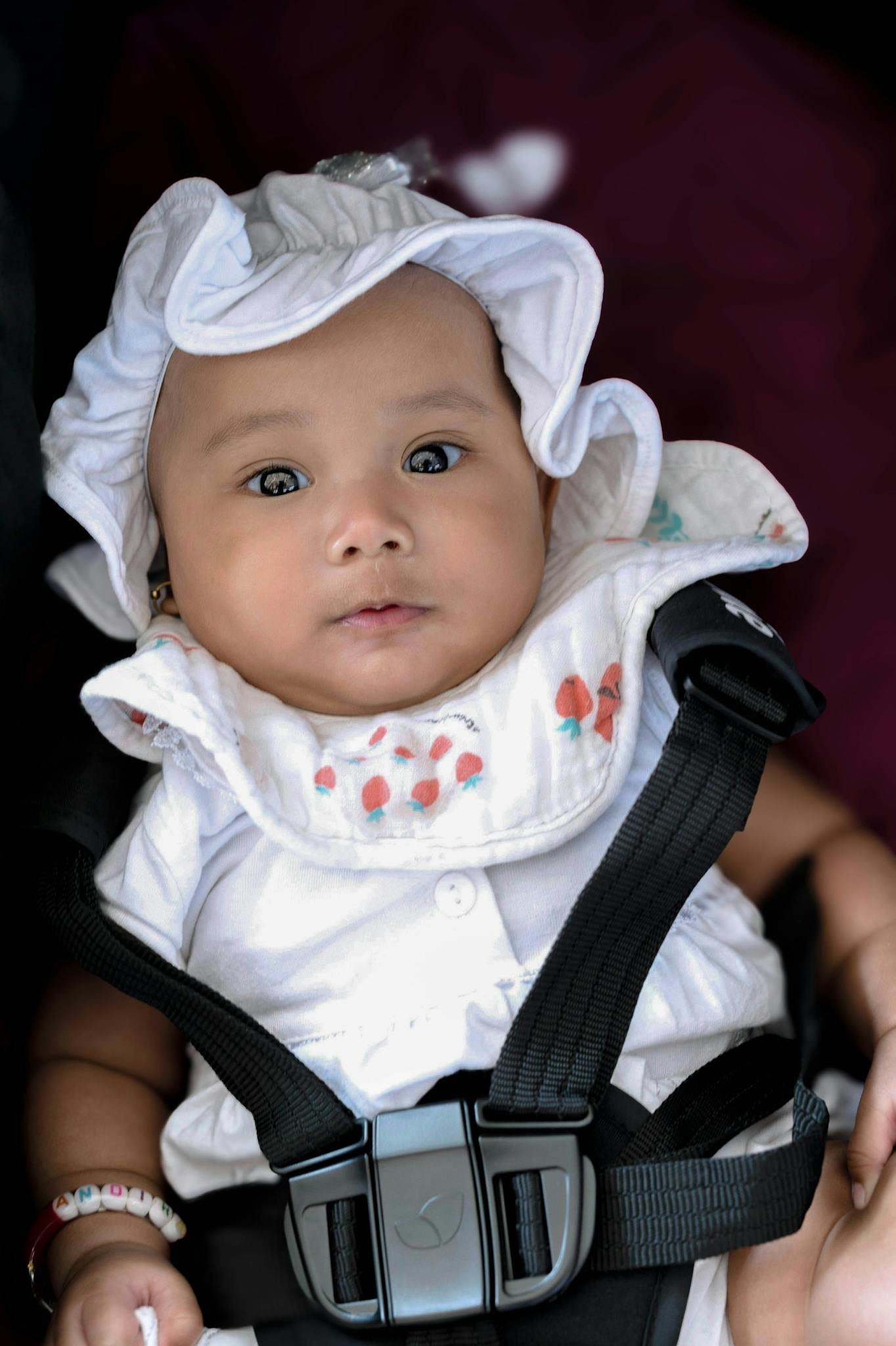 A cute baby wearing a white hat and outfit, safely buckled in a car seat, looking directly at the camera.