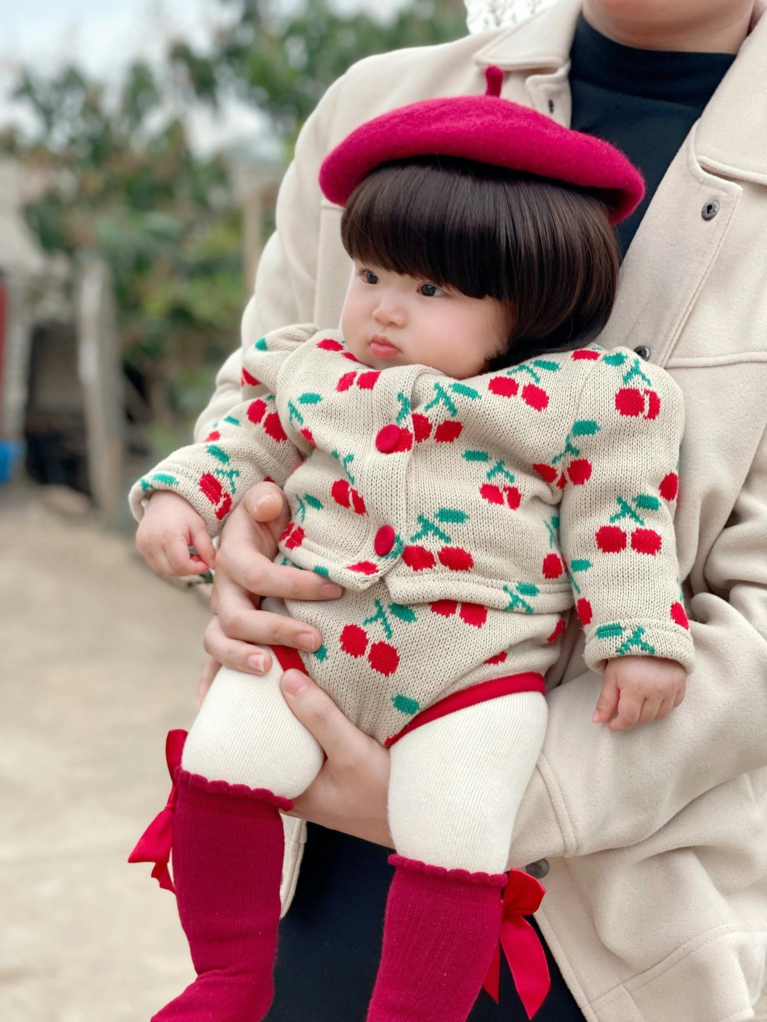 Cute baby in cherry outfit and red beret held by a person outdoors.