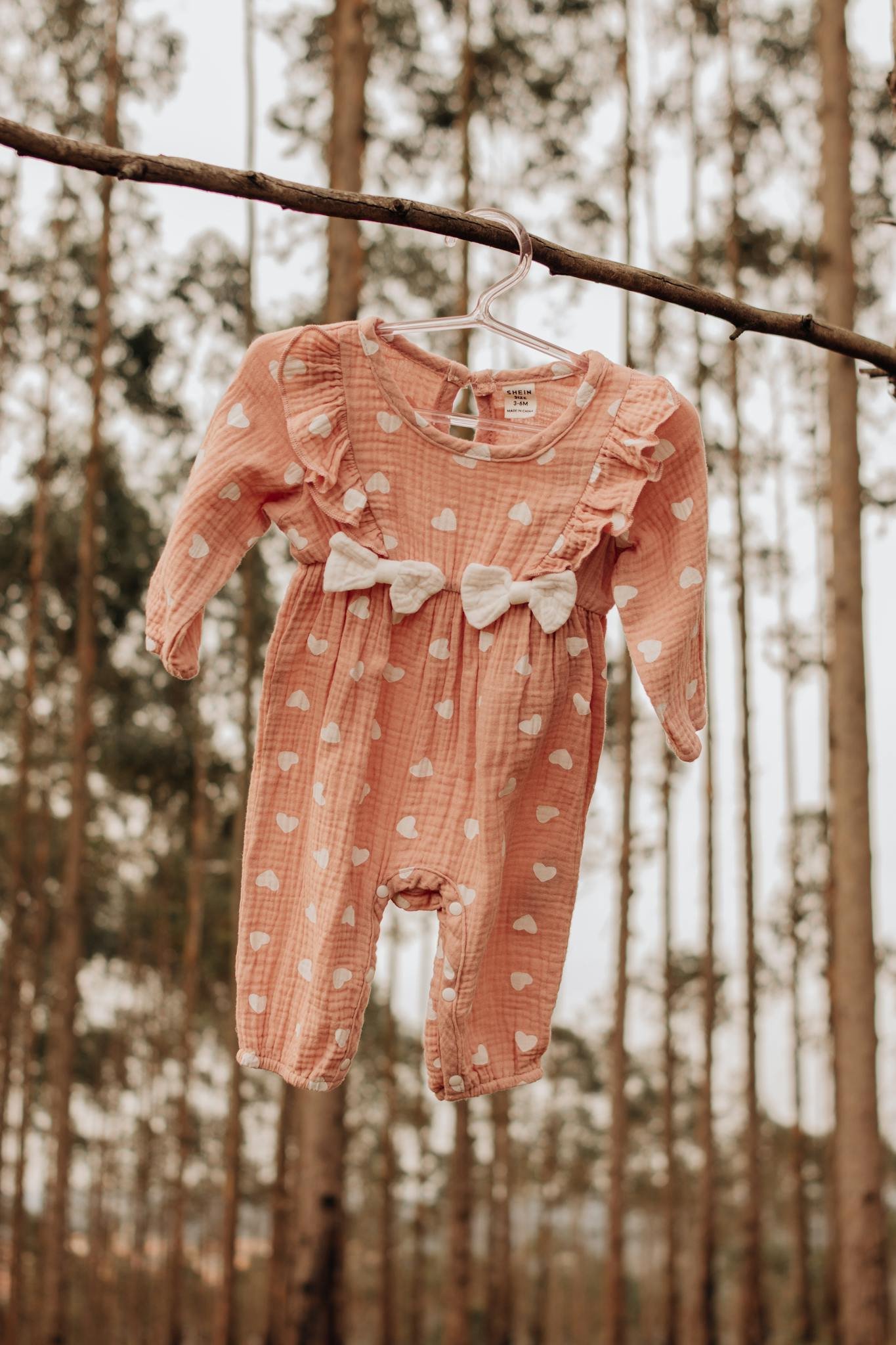 Cute pink baby romper with bows on a hanger in a serene forest setting.